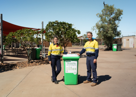 Two people wearing hi-vis clothing are standing beside a green and white Containers for Change bin, at a mine site in the Pilbara.